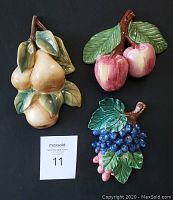 Three ceramic fruit wall decor pieces on black background with auction card. Front view showing coloration and details of pears, apples, grapes.