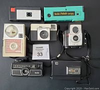 Seven Kodak cameras laid out on a surface showing front views and various designs, colors and conditions.