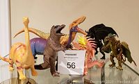 Group photo of 4 dinosaurs and 3 dragons plastic toys arranged in a line on glass shelf.