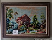 Front view of vintage cross stitch artwork in wooden frame depicting a watermill scene with trees, river and stone bridge.