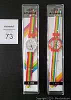 Front view of two Polaroid wrist watches in original clear plastic packaging with rainbow stripe inserts. One watch has a white case, the other red, both with multicolor striped bands.