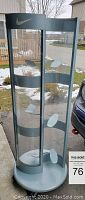 Full view of a cylindrical Nike shoe display stand with glass sides and several interior shoe supports. Shows outdoor background with some snow and pavement.