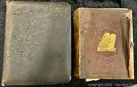 Photo showing the two antique Bibles side by side; the left one has a black leather tooled cover with floral embossing, the right one has a worn brown leather cover with embossed tablets image and significant wear and damage.
