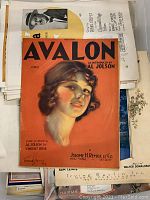 Top view showing 'Avalon' sheet music with a colored illustrated woman's portrait on a red background over various other pieces of vintage sheet music.