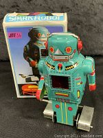 Front angle photo of the light blue wind-up robot with red and yellow accent colors, showing the wind-up key on the side and the metal roller feet, with original box in background.