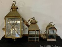 Front view of three vintage electric lanterns on black surface, showing entire group with metal frames and glass panes, varying size from 24", 14" to 11" tall