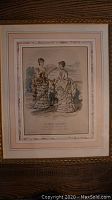 View of first framed hand-colored lithographic fashion print dated 1880, titled La Mode Illustree Paris, showing two women in ruffled period dresses within layered mats and gilded frame.
