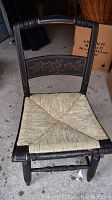Full view of one black wooden rush seat chair with faded gold leaf floral and lyre tole painting on backrest