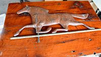 Copper weather vane shaped as a trotting horse with visible green patina, laid on wood surface with mounting rod.