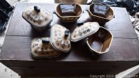 Four hexagonal pottery individual serving dishes with lids displayed on wooden surface. Beige color with blue Chinese characters painted and black knob handles on lids.