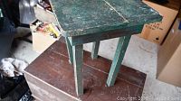 Front view of a green rustic wooden side table showing paint distress and wear, standing on a brown surface.