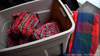 Photo showing several fleece bed sheets or blankets in red and green plaid or checks inside and outside a beige storage bin.