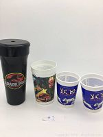 Photo showing the tall black Jurassic Park plastic cup with lid and three other white plastic cups with Jurassic Park and 101 Dalmatians designs.