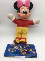 Photo showing full vintage Minnie Mouse plush doll with red shirt, yellow pants, white mittens, pink bow, and red shoes standing behind the 1950's Mickey Mouse blue paint box with colorful Disney character illustrations on the lid.