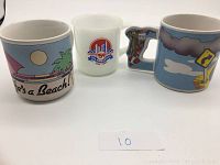 Photo showing all three collectible coffee mugs placed side by side: "Life's a Beach" mug, Toronto 150th Birthday Celebration milk glass mug, and Road Runner mug with character-shaped handle.