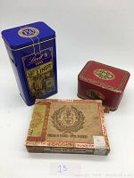 Front view of vintage collectible items including blue Lindt chocolate tin, red McVitie & Price's digestive biscuit tin, and Flor De Manila wooden cigar box