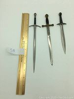 Three sword-shaped metal mail openers shown next to a 12-inch brass ruler for scale.