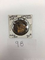 Photo of antique watch fob with Niagara Falls design in a white cardboard holder marked with hand-written details circa 1900 and $30 price