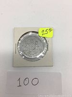 Photo of one metal token in holder with visible coat of arms and inscriptions, price sticker showing $25.00, card with number 100 below
