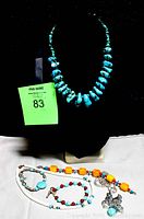 Photo showing a chunky turquoise bead necklace displayed on black bust, with three beaded bracelets/necklaces laid below, including orange, turquoise, and red bead combination with silver charms.
