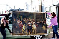 Two people holding the large oil painting outdoors showing full view of historical scene with many figures; shows condition damage such as staple holes and punctures along edges.