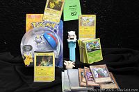 Wide shot showing the M.O.C. Alolan Meowth with Great Ball, banded stacks of Pokemon cards, three Pikachu cards visible, a yellow backed Pokemon card, and scattered Yu-Gi-Oh! cards with brown frames. Pokemon PEZ dispenser in front. Black cloth background.