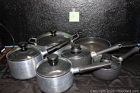 Full set of aluminum pots and pans with glass lids and black handles, arranged on black fabric background