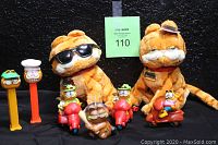 Two Garfield-themed TY Beanie Baby plush toys, one tagged Cool Cat with sunglasses and one Garfield with collar and gold nameplate, surrounded by Garfield PEZ dispensers and PVC figures on scooters
