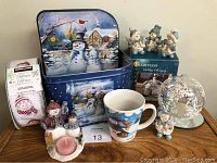 Overall lot image showing all snowman themed items including metal tin, snowman figurines, mug, oil lamp, candle holder, and ribbon roll.