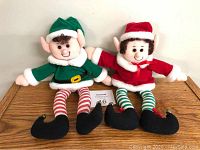 Two plush Christmas elves on a wooden surface, one in green and one in red with striped stockings and curled elf shoes.