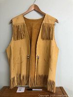 Front view of tan deer skin vest on wooden hanger, fringe along hem, V-neck opening