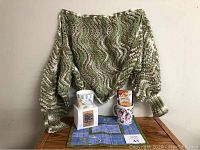 Lot items arranged on wooden surface: hand knit green bed sweater draped in background, ceramic floral mug, boxed scented candle, hot chocolate packet, and decorative fabric mat.