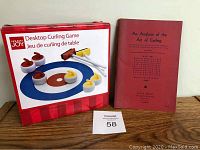 Photo of boxed Holiday Joy Desktop Curling Game and the 1960 softcover book titled "An Analysis of the Art of Curling" by H.E. Weyman on a wooden surface