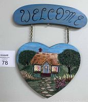 Overall front view of the handcrafted welcome sign with painted cottage and garden on heart shaped wooden board hanging from the blue arched WELCOME top sign.
