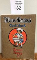 Front cover of the 1915 First Edition Five Roses Cook Book; shows the child chef mixing in a bowl with colorful title text, with visible wear and handwritten name.