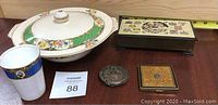 Multiple items on wood background: porcelain covered bowl with floral and green lattice pattern, white cup with blue band and gold detail, vintage compacts and brass box with Nova Scotia emblem visible.