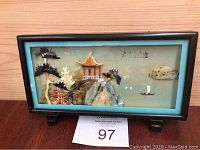 Front view of vintage Asian diorama in wooden frame with glass, depicting pagoda, rocks, foliage, and a sailboat against a blue background.