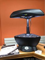 Front view of black AeroGarden SpaceSaver 6 indoor hydroponic garden on a wooden table, grow light panel raised, displaying control buttons on base, with user manual beside it.