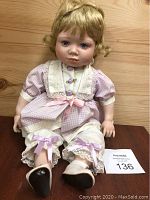 Porcelain toddler doll seated wearing pink gingham dress with lace and bows, shown on a wooden surface.