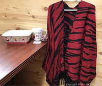 Photo of the reversible red and black cape wrap hanging on a hanger next to a rectangular Christmas Santa Claus melamine bowl, an extra large Christmas themed mug, and a red poinsettia candle on a wooden surface.