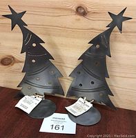 Pair of abstract black metal Christmas tree sculptures with stars on top, displayed side by side on a wooden surface.