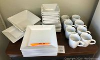View of white modern square-shaped plates, bowls, and mugs arranged on a table showing quantity and style.