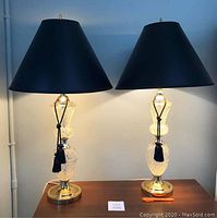 Pair of cut glass table lamps displayed with black lampshades with gold interior and black cord tassels.