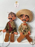 Pair of vintage Mexican marionettes sitting side by side on a white shelf with strings and wooden controls attached, highlighting their ceramic heads, hand painted facial features, and clothing.