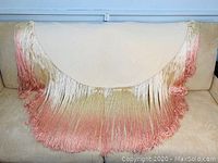 Full view of vintage piano blanket on beige couch, shows semi-circular blanket with long silk tassels in gradient colors from cream to pink.