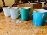 Four Le Creuset ceramic mugs placed in a row on a wooden table in beige, white, turquoise, and blue colors.