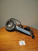 Front and side angled view of the vintage spotlight laying on a wooden table. The spotlight head has a glass lens and the black power cord is coiled around the handle.