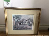 Framed print showing the full piece with beige mat and gold frame. The print depicts a historic village scene with houses, a horse-drawn carriage and figures.