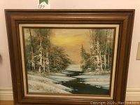 Framed oil painting showing entire artwork of snowy forest and river at sunset with warm sky tones.