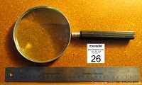 Magnifying glass with metal rim and black handle shown alongside a stainless steel ruler indicating lens diameter.